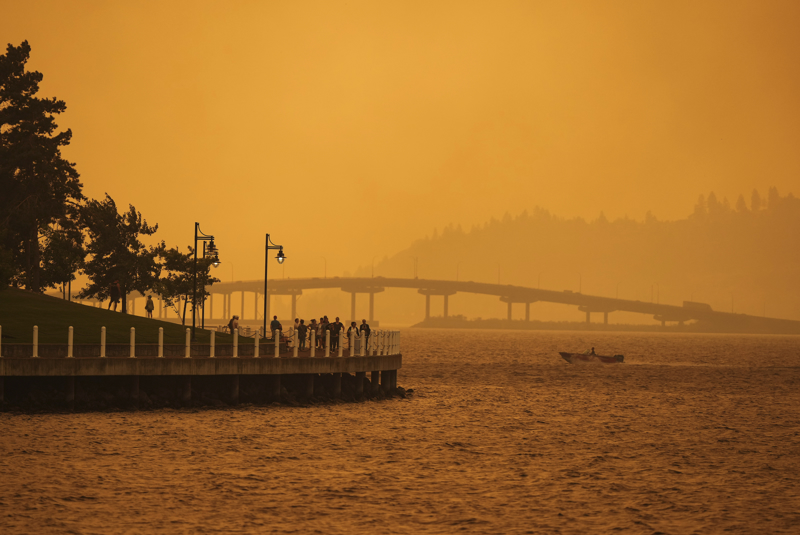 Eine Person fährt in einem Boot an Menschen vorbei, die auf der Promenade spazieren gehen, während der Rauch des McDougall Creek Waldbrandes das Gebiet am Okanagan Lake in Kelowna, British Columbia, bedeckt.