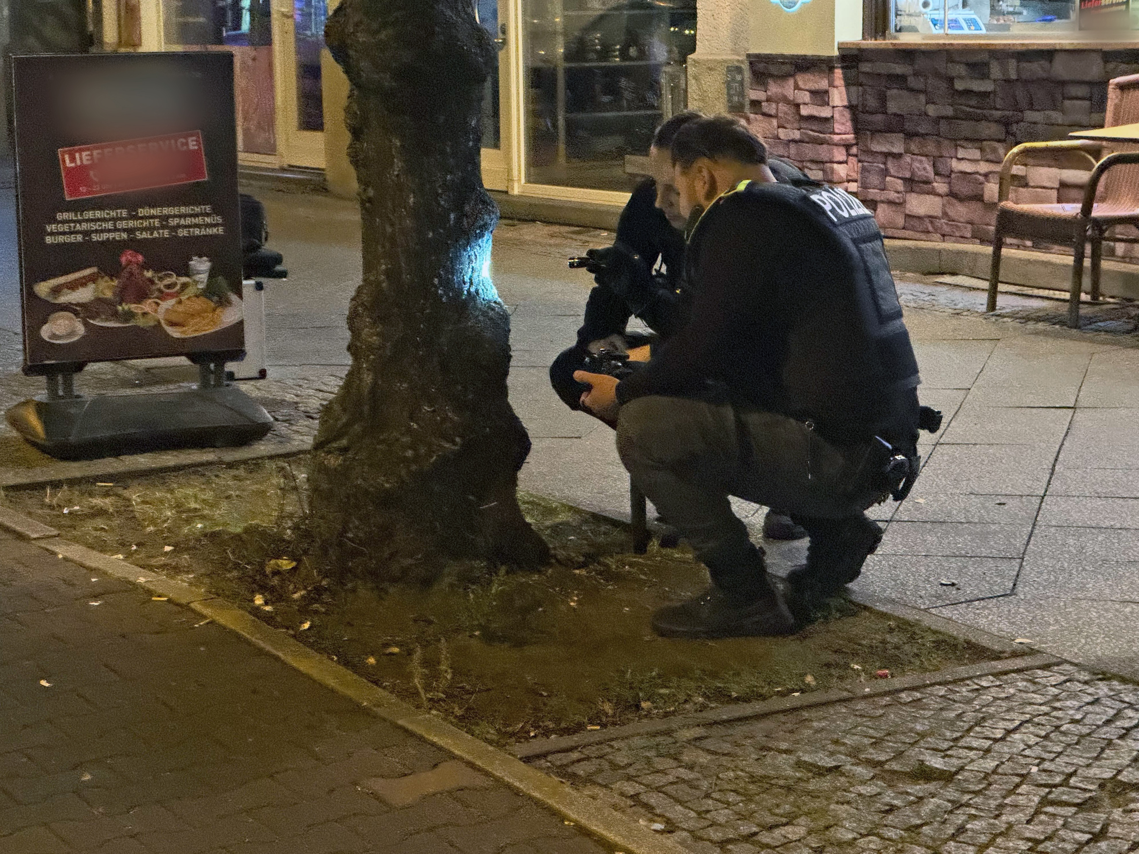 Montagabend in Berlin-Reinickendorf: Zwei Polizisten begutachten eine Spur, die eine Kugel an einem Baum hinterlassen hat. Zuvor ist ein Mann an einem Imbiss angeschossen worden.
